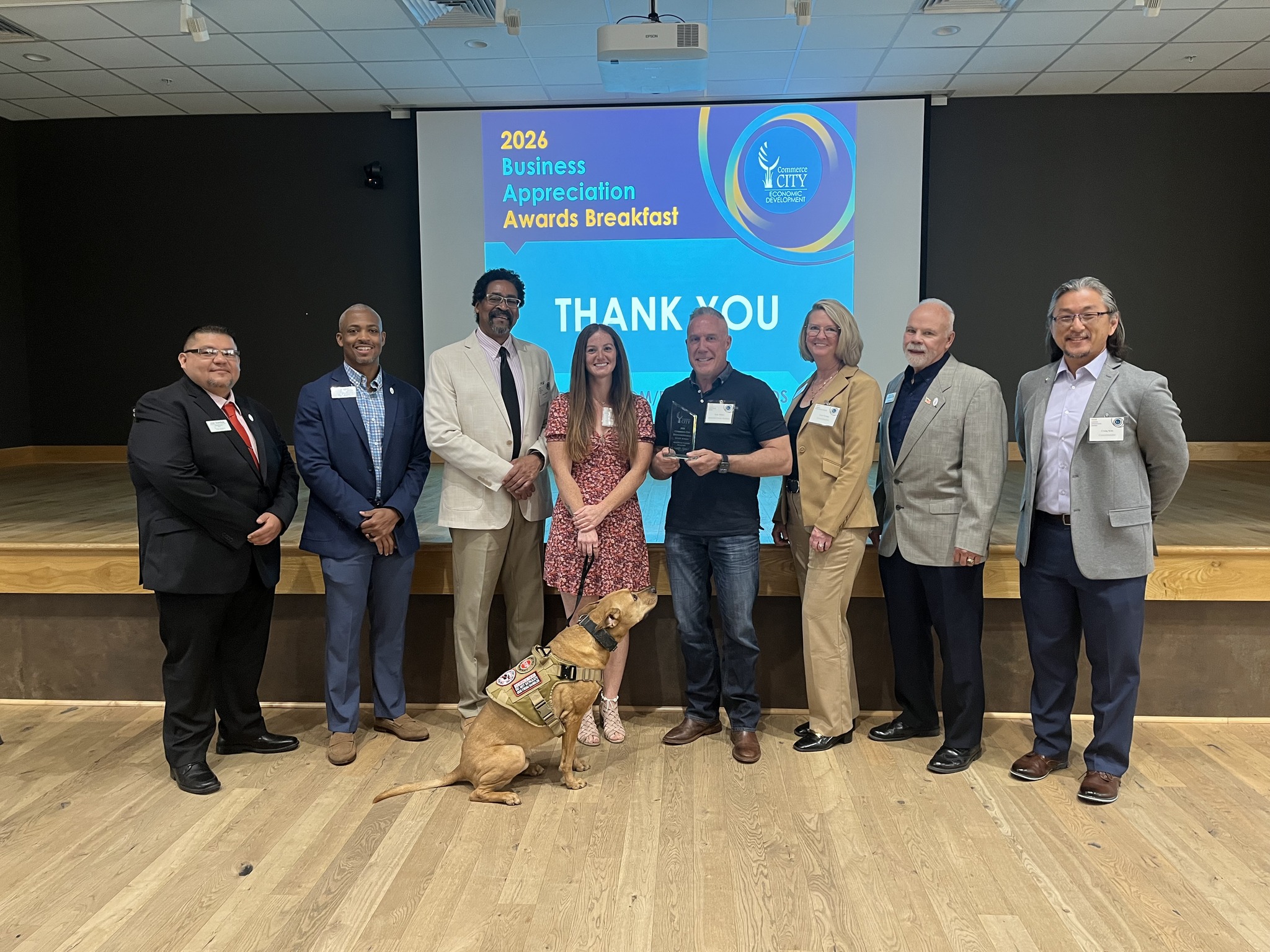 Group of eight adults in business attire posing on a stage with a service dog at the front center.